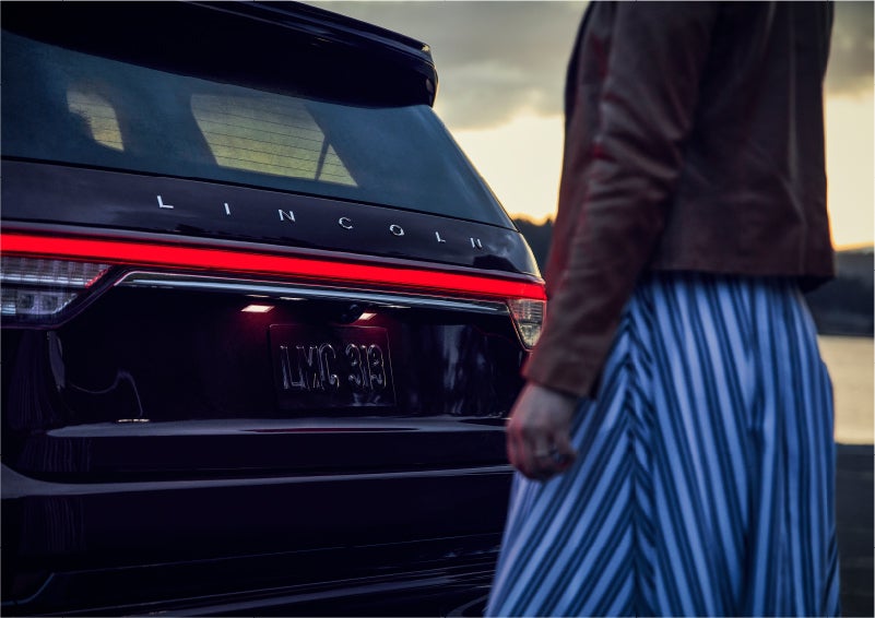 A person is shown near the rear of a 2023 Lincoln Aviator® SUV as the Lincoln Embrace illuminates the rear lights | Napleton River Oaks Lincoln in Calumet City IL